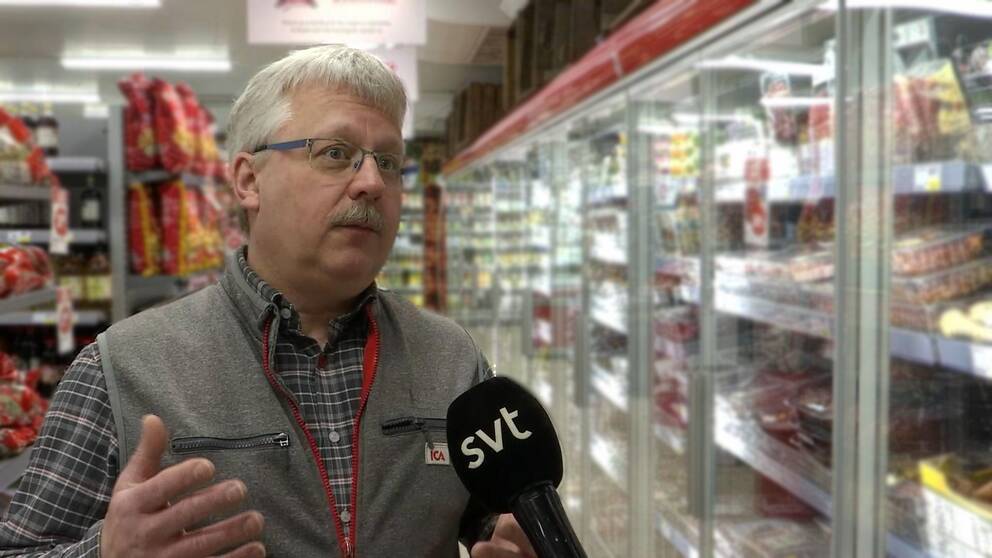 Man med mustasch och glasögon står i en matbutik med en mikrofon framför ansiktet.