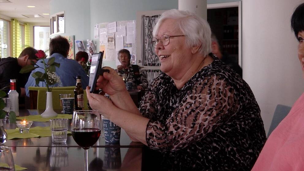 En kvinna skrattar och visar upp något på sin mobil för en vän. Hon sitter vid ett bord med vinglas och chips framför sig.