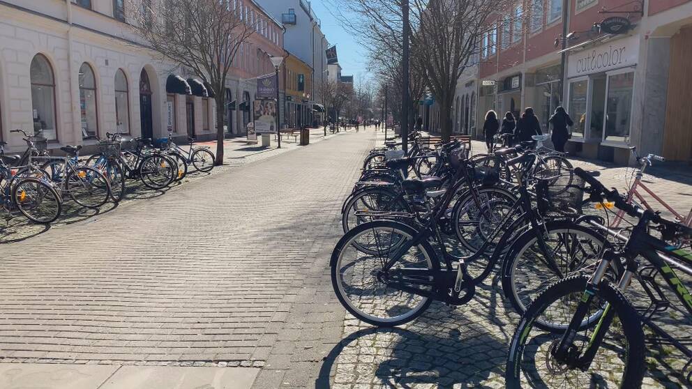 En folktom gågata med cyklar, parkerade i cykelställ, i förgrunden.