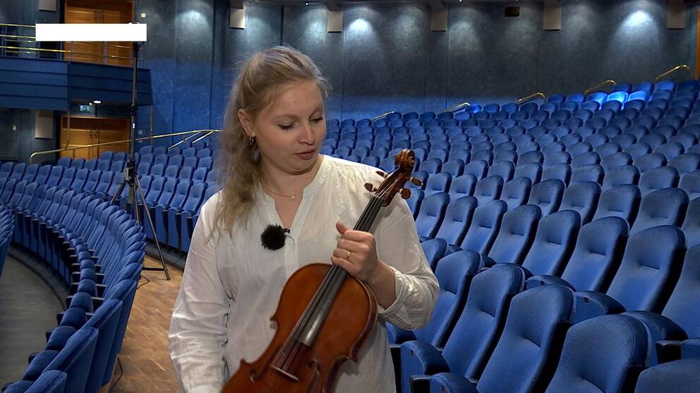 Jenny håller i sin viola, en altfiol. Bakom henne tomma publikstolar i den blå konsertsalen i De Geer-hallen i Norrköping.