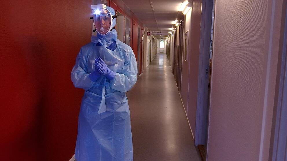 Therese står med blå handskar, blå plastöverdragskläder, blå huva och en mask med genomskinklig plast över ansiktet och ovanför sitter en vit liten dosa som är en fläkt med en liten lampa i. Hon står i en lång korridor.
