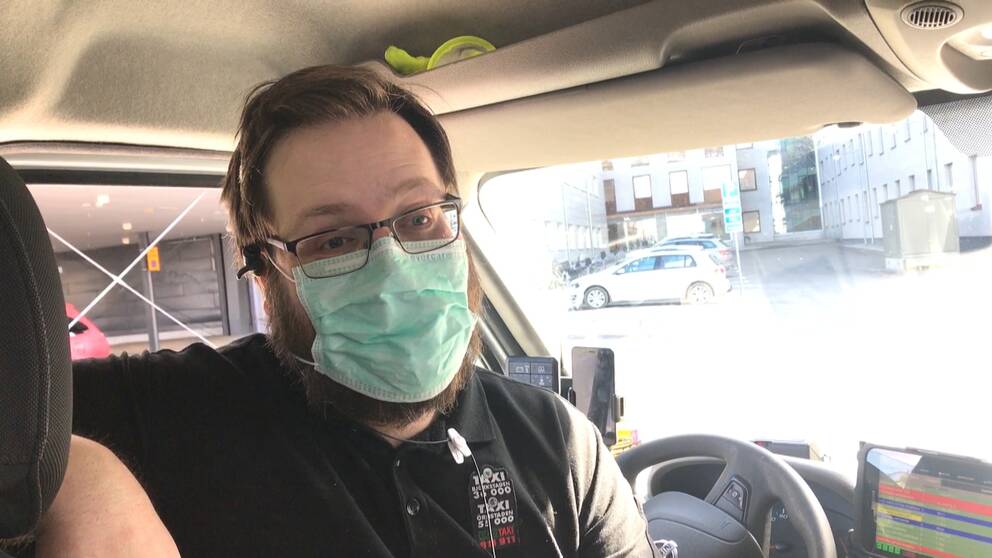 En taxichaufför med glasögon och munskydd som sitter vid ratten i sin bil utanför Norrlands universitetssjukhus i Umeå. Han har svart pikétröja.