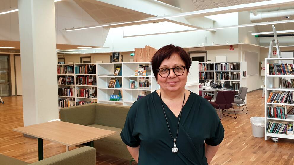 En kvinna med kort mörkbrunt hår, glasögon och grön tröja tittar in i kameran. Hon står i ett skolbibliotek.