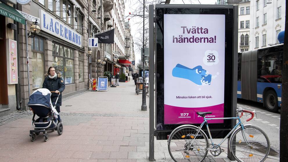 En kvinna med barnvagn passerar en annonsskylt med en annons från Folkhälsomyndigheten. ”Tvätta händerna” står det i annonsen.