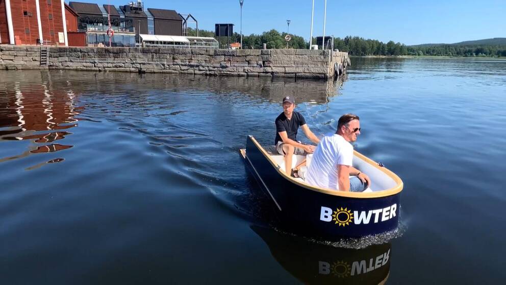 Två män sitter i en liten elbåt, en i fören och en styr i akten, på väg ut från en hamnpir i sten som syns i i bakgrunden.