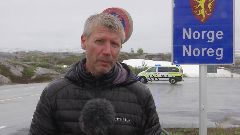 Reportern står vid gränsen till Norge. Hans står bredvid gränsskylten till Norge och i bakgrunden syns en norsk polisbil.