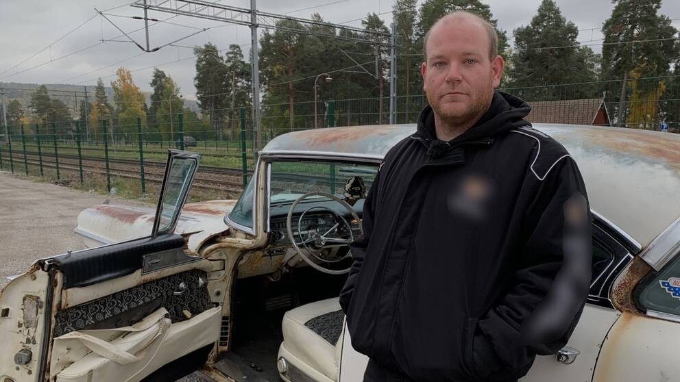 en snaggad man star lutat mot en raggarbil i Rättvik. Bilen är vit och rostig. Mannens jacka är svart.