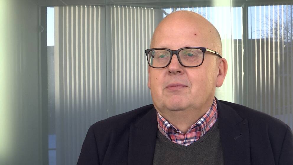 Skallig man med glasögon. Mannen har mörk kavaj, v-ringad grå tröja och en rödvitblårutig skjorta.