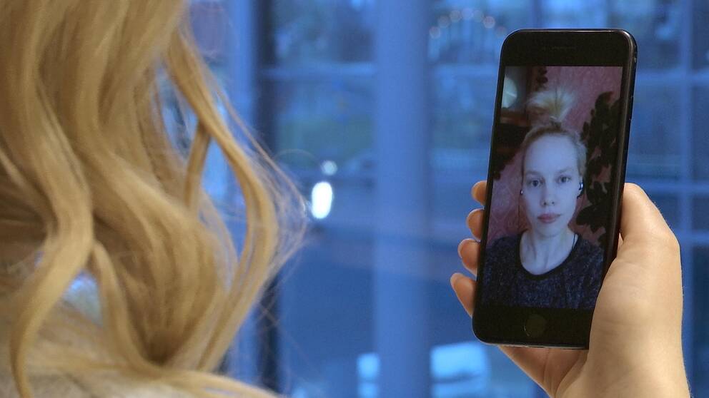 En kvinna med blont hår uppsatt i tofs syns på en mobilskärm. Hon har svart tröja och hörlurar i öronen.