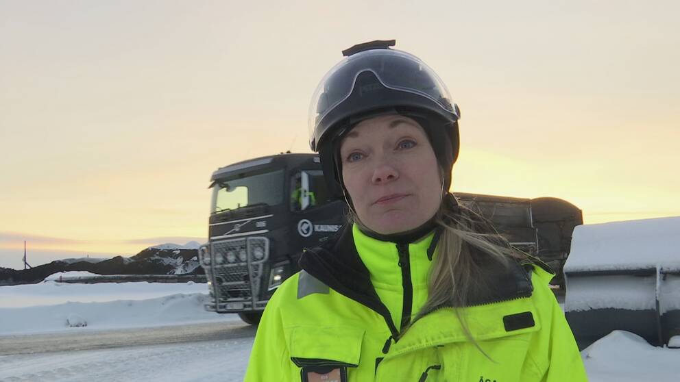 en kvinna i hjälm och varselkläder utomhus vintertid, lastbil i bakgrunden