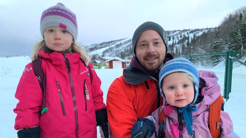 Två mindre barn i färgglada overaller bredvid en vuxen man som hukar sig ner. Alla tittar in i kameran.