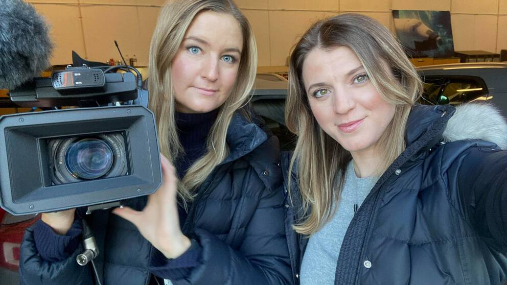 Två kvinnor med blondt hår och blåa jackor. I ena handen hålls en videokamera.