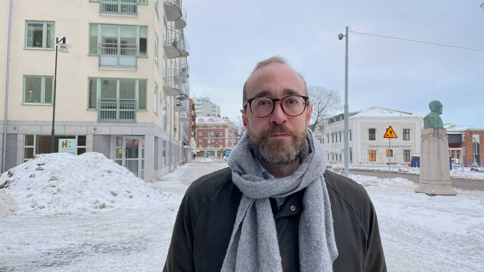 NÄRBILD PÅ MEDELÅLDERSMAN SOM TITTAR IN I KAMERAN. SNÖ, HUS OCH BUSSHÅLLPLATS I BAKGRUNDEN.