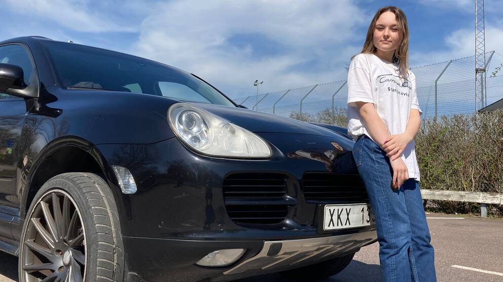 En ung tjej i 15-årsåldern står och lutar sig mot motorhuven på sin Porsche Cayenne som är ombyggd till en A-traktor. 