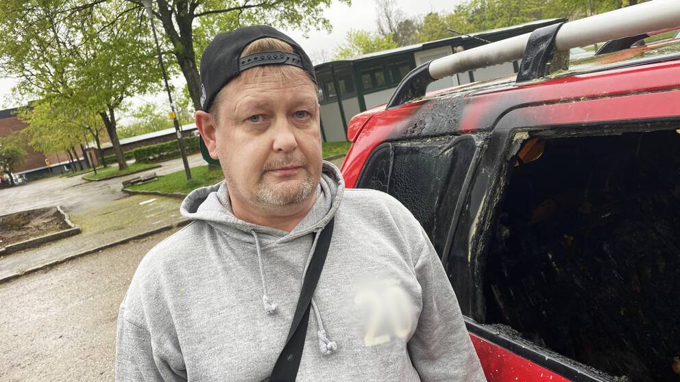 Lars står intill sin utrbrända bil på en parkeringsplats.