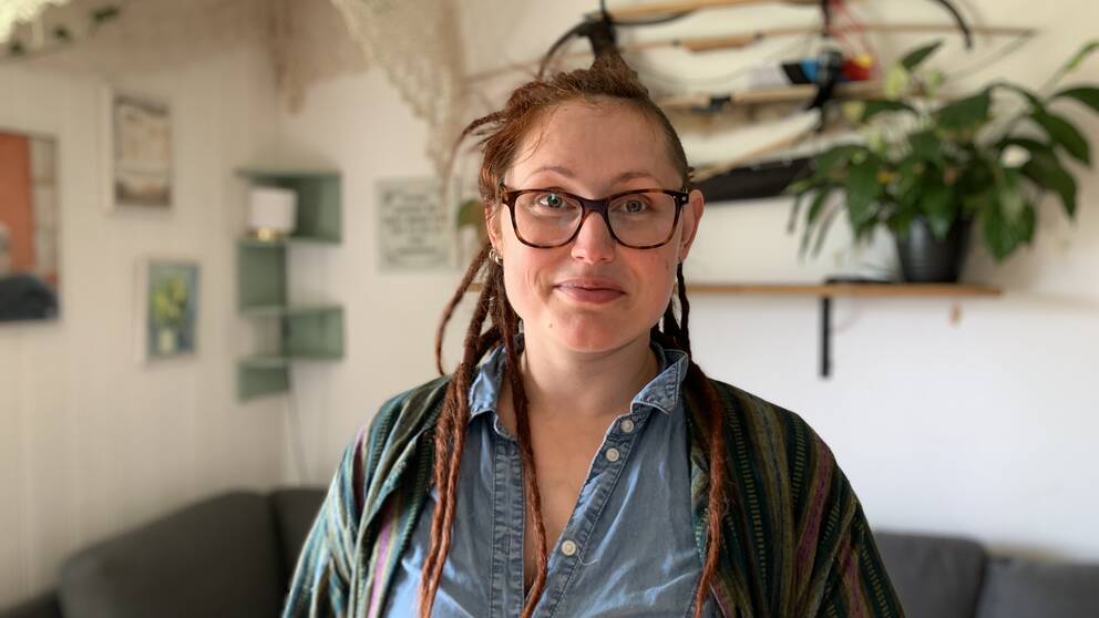 Tanja Östberg har röda dreadlocks och glasögon. Tanja har en jeanblus och en cardigan på sig. Hon står i sitt vardagsrum framför soffan och poserar. I bakgrunden hänger några pilbågar på väggen.