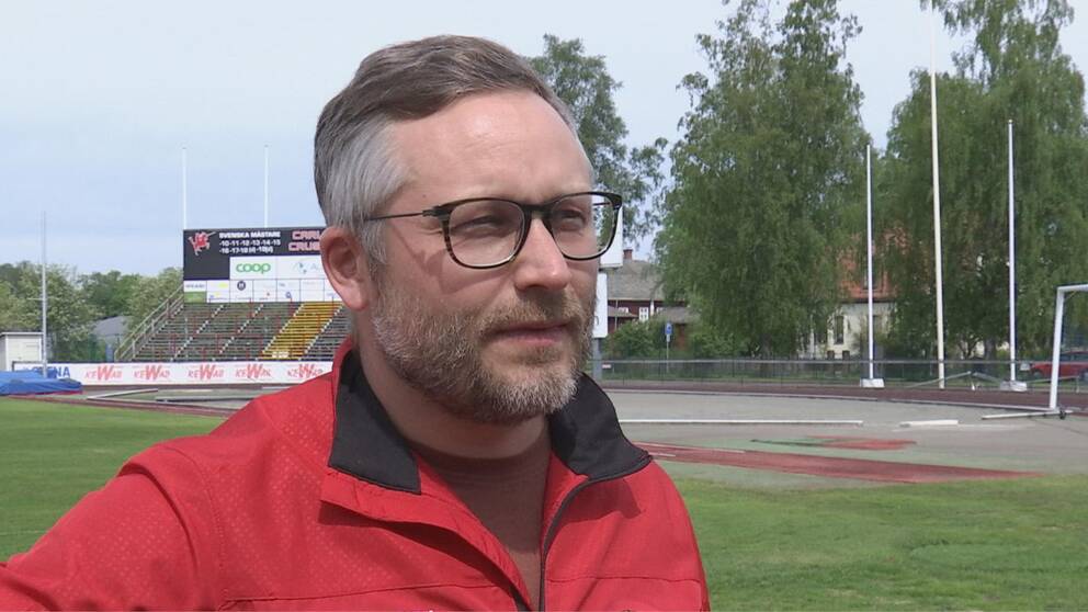 Man i glasögon och kort skägg, röd jacka står på en friidrottsarena.