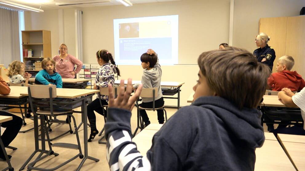 En elev som räcker upp handen, i bakgrunden syns andra elever och en lärare.