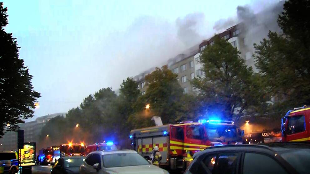 Rök kommer ut ut ett stort flerfamiljshus. Räddningstjänstens bilar står i förgrunden.