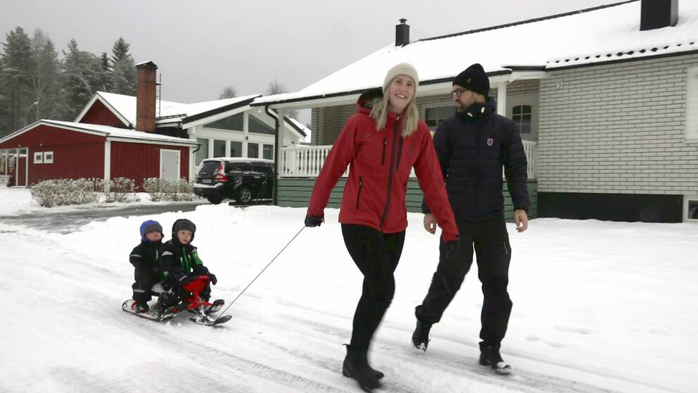 En man och en kvinna går på en snöig väg. Kvinnan drar två små barn på en bob. I bakgrunden syns villor.