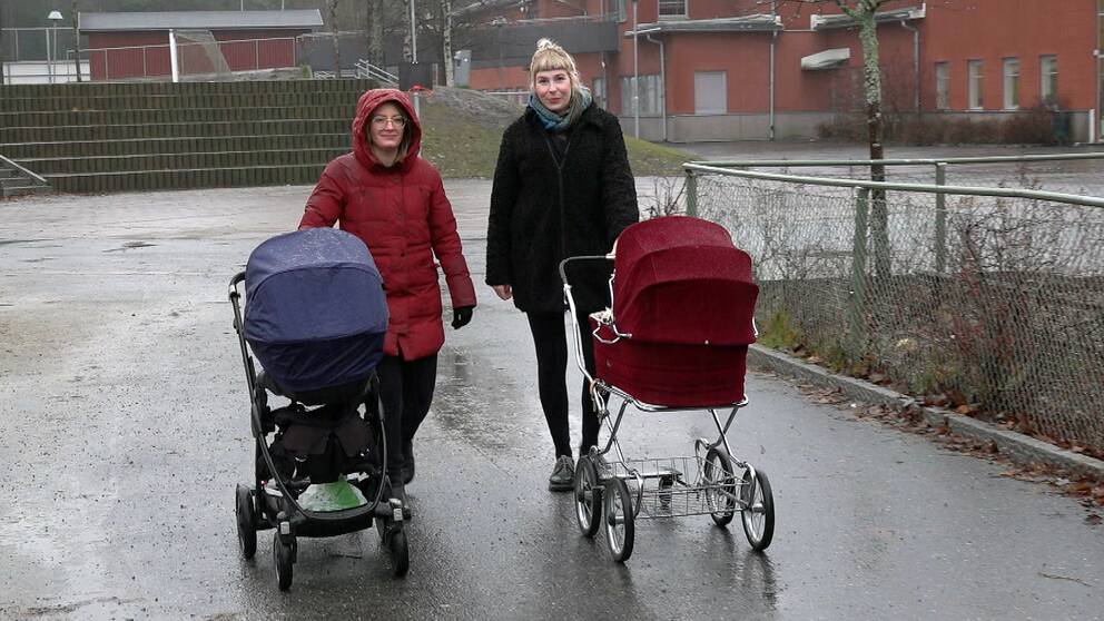 Två kvinnor med varsin barnvagn står på en regnig asfaltsyta. 