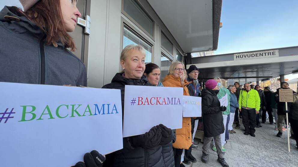 Flera vinterklädda personer håller upp skyltar med olika budskap vid sjukhuset.