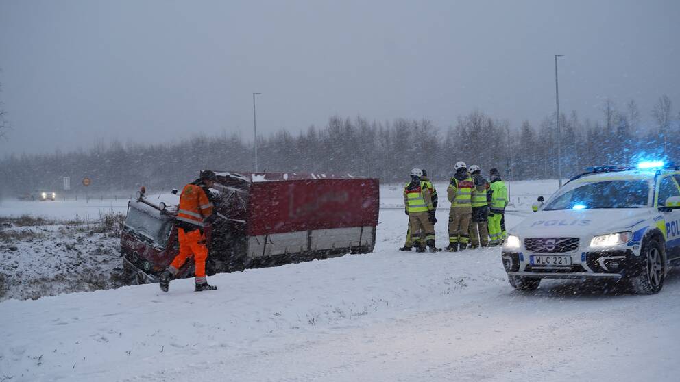 Bild på en trafikolycka. En lastbil har kört av vägen och står på alla fyra däck i ett dike. Det snöar. Med på bilden är bärgaren, räddningstjänsten samt en polisbil. 