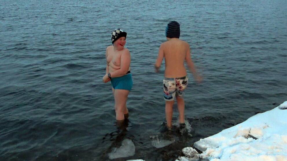 två pojkar i badbyxor och mössa kliver i vattnet från en snökant.