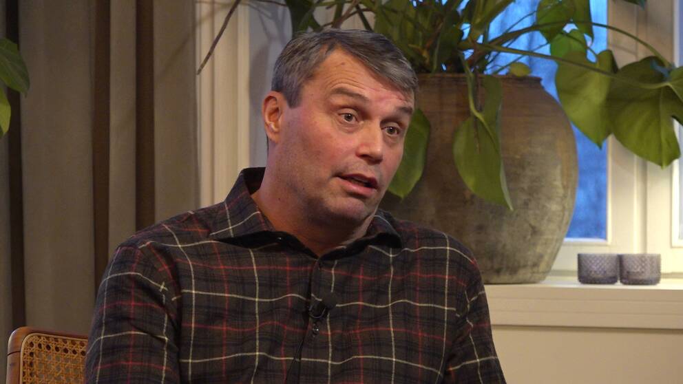 Daniel  Kindberg iklädd rutig skjorta sitter i sitt vardagsrum.