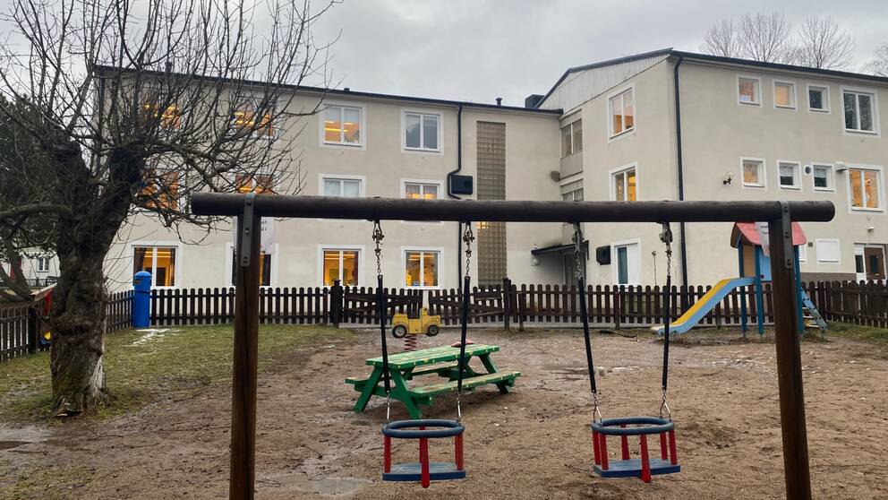 Bild på Ålidenskolan och Ålidens förskola i Flen. I förgrunden syns en utegård med gungställning, rutschkana och en grön träbänk.