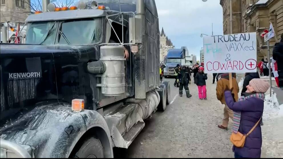 Två lastbilar står parkerade på en gata. En kvinna håller upp protestskylt.