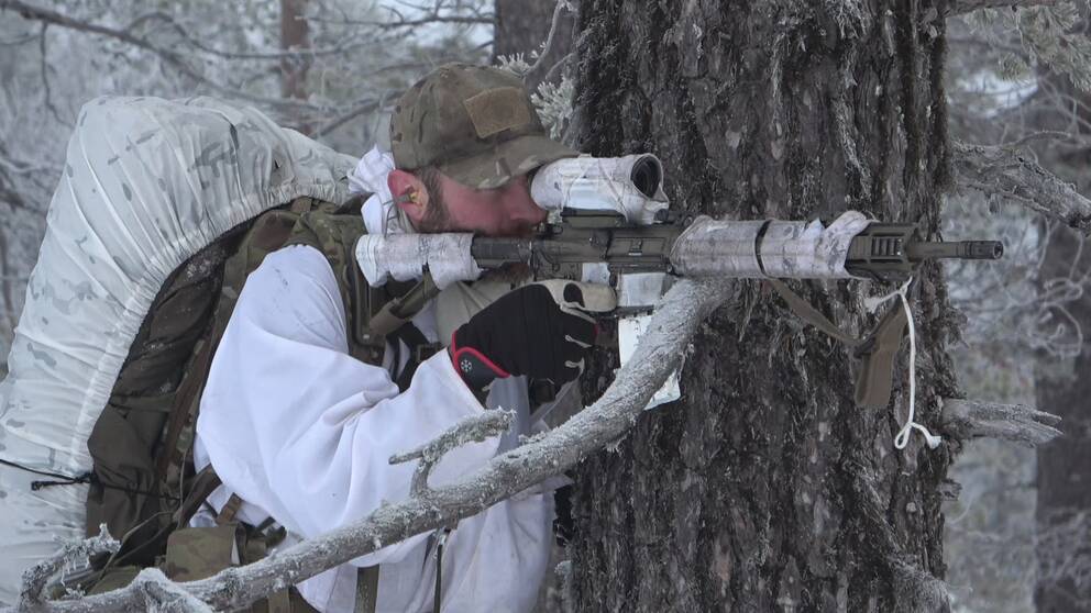 Jonas Dahl, en soldat i vinterkamouflage med stor ryggsäck siktar med sitt vapen intill ett träd