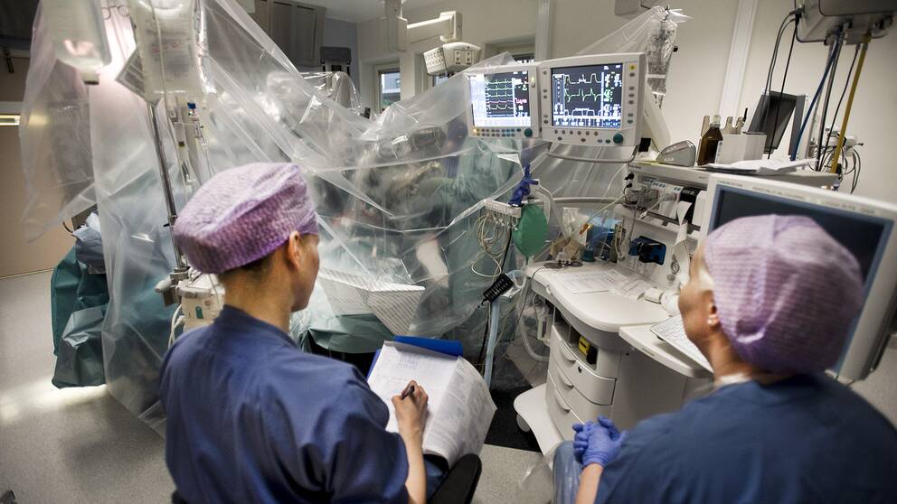 Vårdpersonal i operationskläder sitter bredvid en pågående operation i en sjukvårdssäng. 