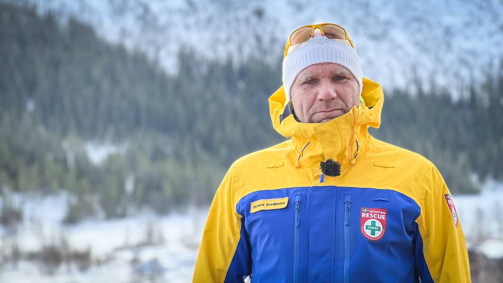 En man i blågul skidjacka ser in i kameran med fjällmiljö i bakgrunden.