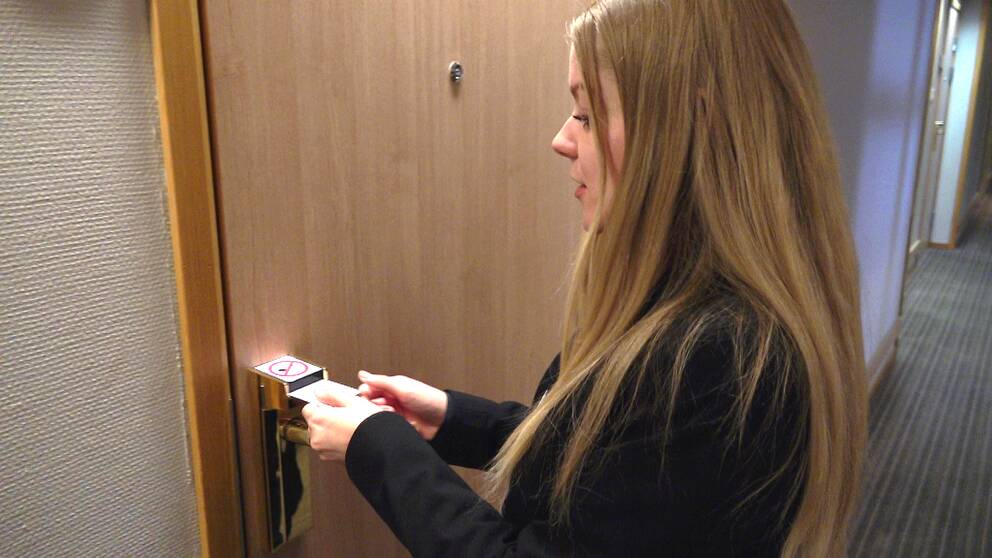 En ung blond kvinna med svarta kläder håller fram ett hotellkort mot en stängd dörr.