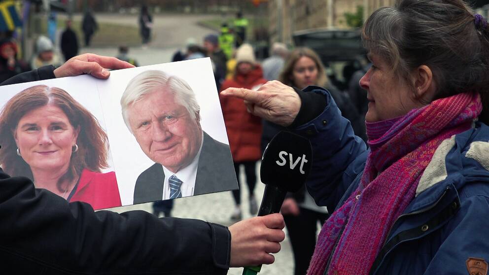 En kvinna pekar på en bild på Johnny Magnusson (M) och Helén Eliasson (S), som en reporter håller upp. Hon befinner sig på en gata i Haga med folk gåendes bakom henne.