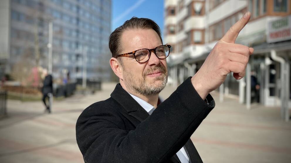 En man med glasögon som pekar på bostadshus i en förort.