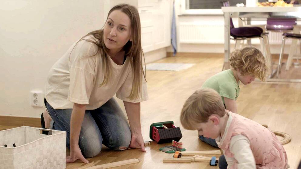 En kvinna sitter på golvet bredvid två mindre barn som leker med en tågbana i trä. 