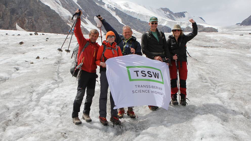 Fem personer iklädda friluftskläder håller en flagga med texten ”Transibirian Science Highway”