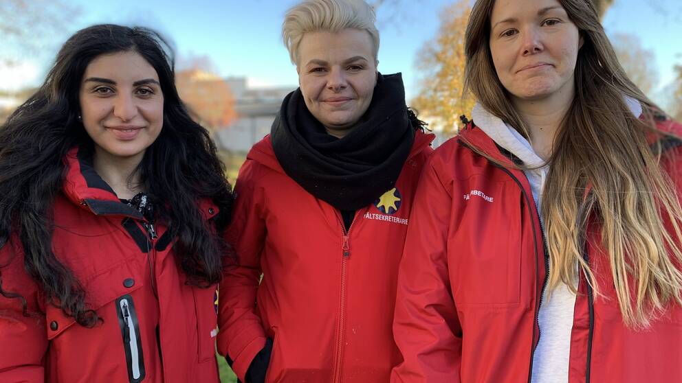 Porträttbild på de tre samordnarna som står i röda jackor med träd bakom.