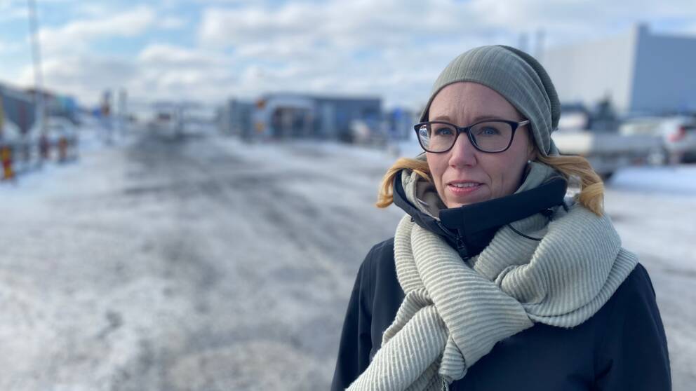 En kvinna i glasögon, halsduk och mössa, framför den nya batterifabriken i Skellefteå