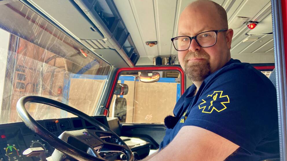 en man med glasögon klädd i tshirt med ambulanssjukvårds-märke på ärmen sitter i hytten på ett blåljusfordon, tittar mot kameran