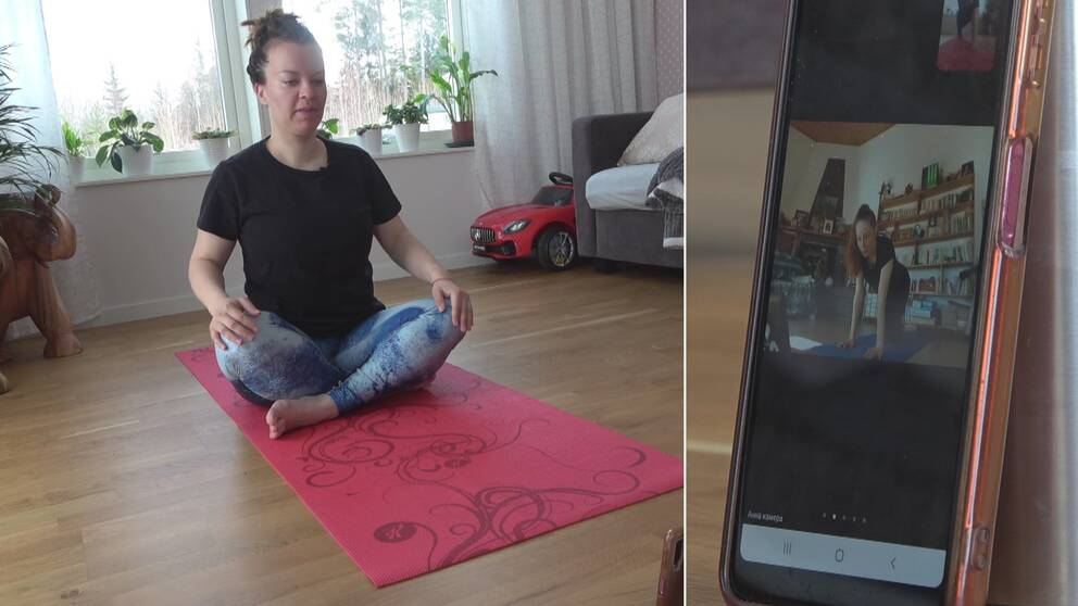 En kvinna i yoga-tights sitter i med benen i kors på en yoga-matta. En yogainstruktör syns på en mobilskärm.