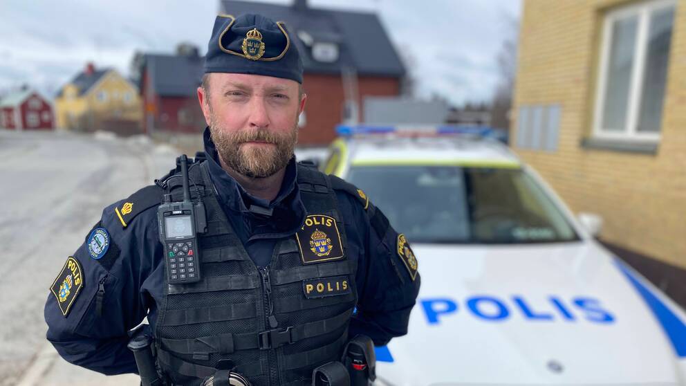 en man med skägg, i polisuniform, framför en polisbil i ett villaområde