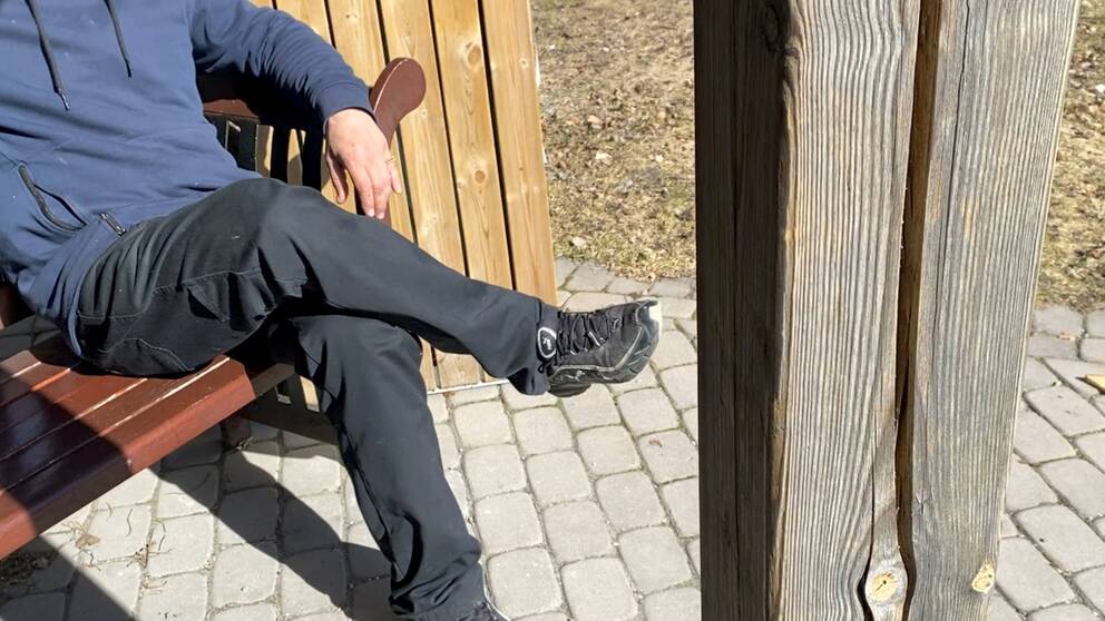 person vars ansikte är utanför bild, sitter på en bänk på en uteplats