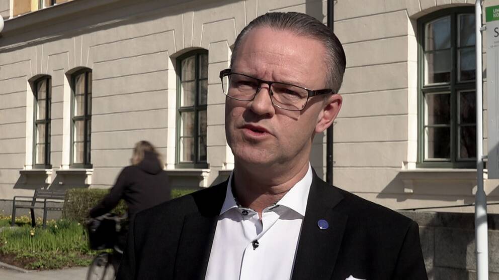 En man i kavaj och klasögon som står framför ett hus.