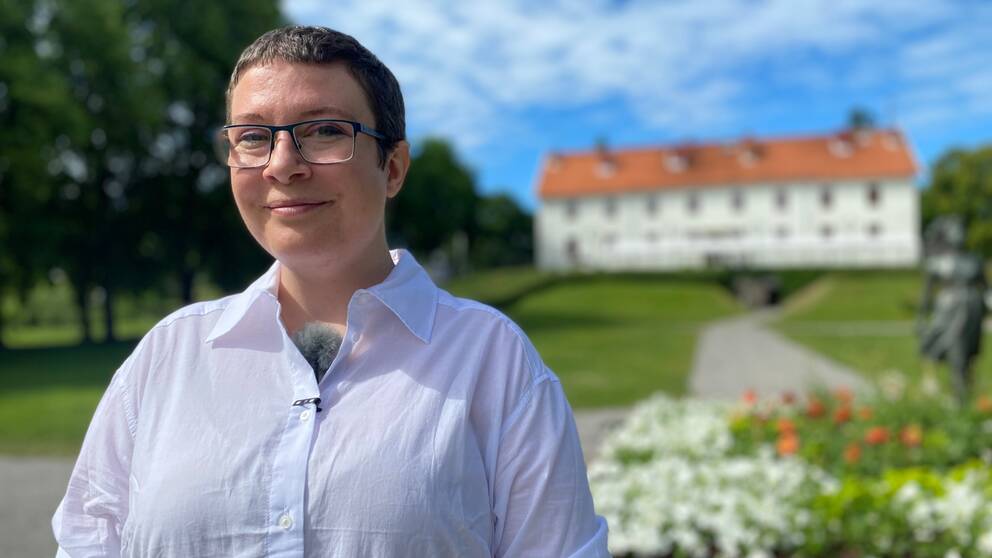 Bild på antikvarien, Cecilia Bergman. Hon tittar in i kameran och ler. I bakgrunden syns en grön gräsmatta, blommor och Sundbyholms slott. 