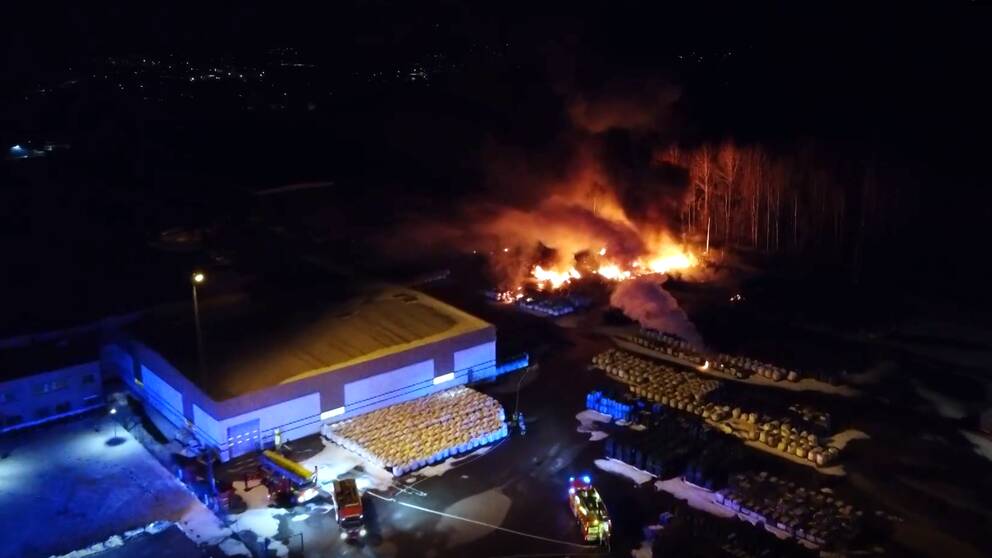Vy från ovan i natt, över stor eldhärd bredvid lagerbyggnad. Två brandbilar i nederkant av bilden.