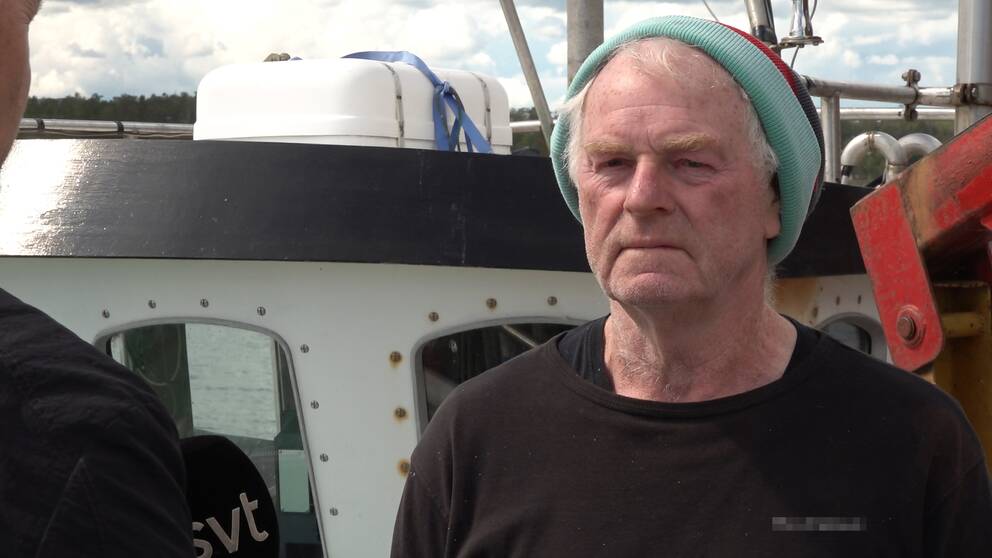 närbild, en äldre medelåldersman i tröja och mössa intervjuas på en fiskebåt