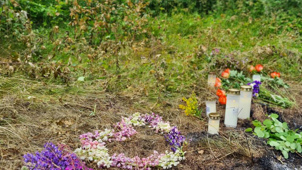 blonkrans och gravljus på marken vid en förkolnad fläck, skog bakom